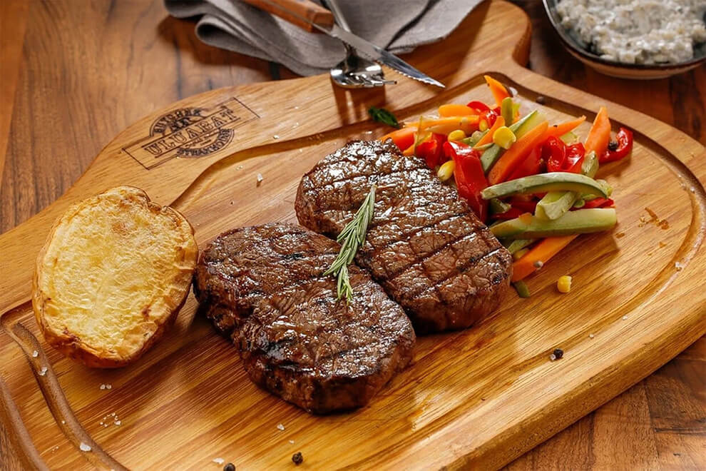 Zwei gegrillte Steaks auf einer Holzplatte angerichtet mit rotem Paprika und Weissbrot.